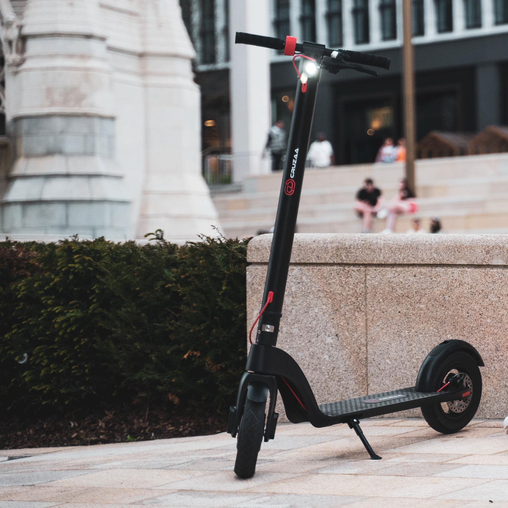 The Commuta Scooter - In its standing position and with the full beam on. It is photographed outside in an urban setting. 