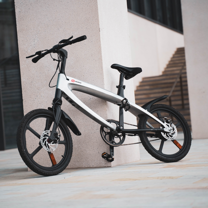 The Cruzaa E-Bike in Gunmetal Grey, in an urban setting stood up against a pillar. 